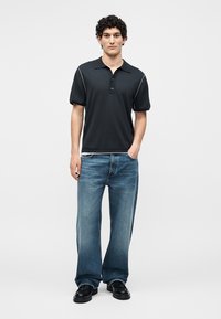 Jeune homme aux cheveux bouclés portant un polo noir, un jean bleu à jambes larges et des chaussures noires, debout avec une main dans la poche.