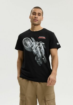 Mężczyzna ubrany w czarną koszulkę z grafiką motocrossowca i beżowe spodnie cargo, stojący na jasnym, jednolitym tle.