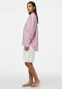Camicia oversize a strisce rosse e bianche con taschino sul petto, abbinata a una gonna bianca a ginocchio e infradito, mostrata di lato.