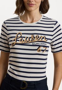 Dryžuota medvilninė marškinėliai kreminės ir tamsiai mėlynos spalvos, su auksiniu siuvinėtu "Lauren" logotipu ir "Ralph Lauren 67" detalėmis. Apvalus kaklo išsikirpimas.