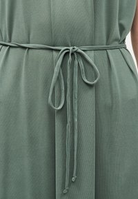 Φόρεμα από απαλό πράσινο ύφασμα με λεπτή ζώνη δεμένη σε απλό φιόγκο στη μέση, με ελαφριές πτυχώσεις στο ύφασμα.