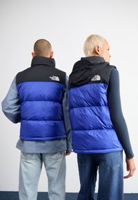 Twee personen dragen gewatteerde vesten, de ene in het blauw met een zwarte bovenkant, de andere in grijs. Beide hebben capuchons en merkvermelding op de achterkant.