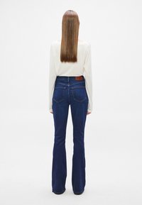 Μπλε τζιν καμπάνα με ψηλή μέση, δύο πίσω τσέπες και δερμάτινη ετικέτα. Λείο ύφασμα και πλούσιο χρώμα που αναδεικνύουν το σχέδιο.