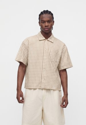 Homme aux cheveux tressés portant une chemise beige à carreaux à manches courtes et un short écru, debout devant un fond blanc uni.
