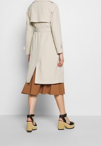 Béžový trenčkot s páskem v pase, bočním rozparkem a zapínáním na knoflíky u manžet. Kombinován s hnědou rozšířenou sukní a černými sandály na platformě.