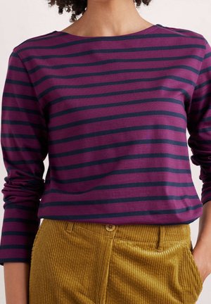 Vrouw draagt een langarmig paars-zwart horizontaal gestreept shirt, ingestopt in mosterdgele corduroy broek met knoop en zak.