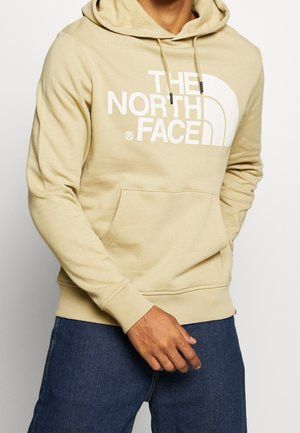 Beige hættetrøje med frontlomme og hvidt "THE NORTH FACE" logo, båret med blå denimjeans, mands overkrop og hånd synlige.