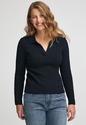 Marineblaues Kabelstrickpullover mit geripptem Saum und Bündchen, V-Ausschnitt und strukturierten Musterdetails. Kombiniert mit blauen Jeans.