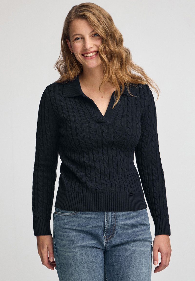 Granatowy sweter z dzianiny w warkocze, z prążkowanym dołem i mankietami, z dekoltem w kształcie litery V oraz detalami o fakturowanym wzorze. W zestawie z niebieskimi dżinsami.