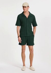 Dunkelgrünes, kurzärmeliges, geripptes Hemd mit Kragen, kombiniert mit passenden Shorts. Beinhaltet eine beige Mütze und cremefarbene Slip-On Schuhe.