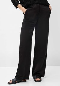 Pantalon noir à jambe large en matériau lisse avec poches latérales. Associé à des sandales noires plates à brides croisées. Design simple et minimaliste.