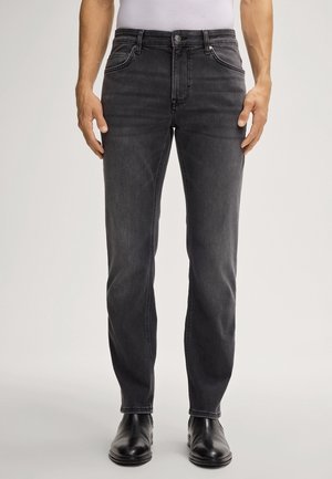 Homme portant un jean noir slim délavé avec poches avant et des bottes en cuir noires, debout devant un fond clair uni.