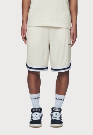 Beżowe spodenki do koszykówki z czarno-białym paskowanym wykończeniem, noszone z białymi skarpetkami i czarnymi zamszowymi trampkami z białymi akcentami.