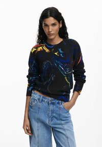 Pull en tricot noir avec des motifs abstraits multicolores en bleu, rouge et jaune. Encolure ras du cou et manches longues. Assorti à un jean bleu clair.