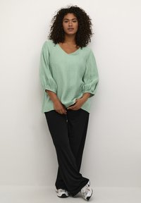 Blusa verde menta con texture, scollatura a V e maniche a palloncino, abbinata a pantaloni neri larghi e scarpe sportive grigio e bianche.