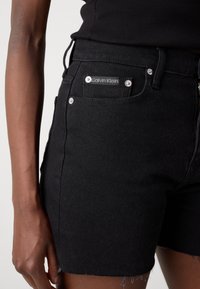 Black denim shorts with a high waist, featuring a silver Calvin Klein logo and metal hardware. The fabric has a slightly textured finish.