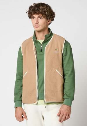 Jeune homme portant un gilet en polaire beige par-dessus un pull à fermeture éclair quart de tour vert et un pantalon blanc, debout devant un fond uni.