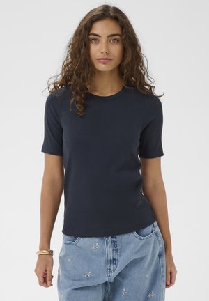 Ženska z rjavimi kodrastimi lasmi, oblečena v mornarsko modro majico s kratkimi rokavi in svetlo modre kavbojke z drobnim cvetličnim vezenjem, stoji pred belim ozadjem.