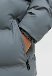 Grå pufferjacka i mjukt material, med en dragkedjeficka på ärmen och samlade manschetter för en tät passform.