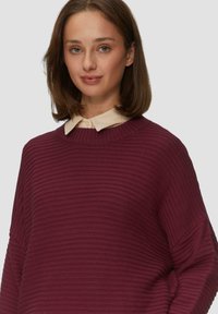 Pull en crochet bordeaux avec un col rond et de larges manches, superposé sur une chemise beige à col. Texture douce et coupe décontractée.