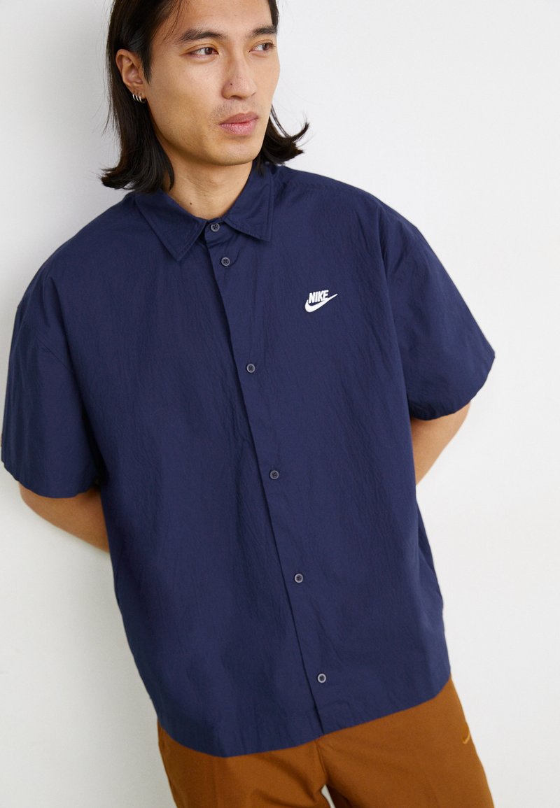 Chemise à manches courtes bleu marine avec une finition texturée, dotée d'une fermeture à boutons et d'un petit logo Nike blanc sur la poitrine gauche.
