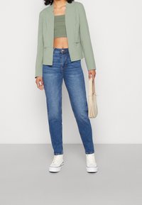 Blazer cropped vert clair au design structuré, associé à un jean bleu taille haute et des baskets blanches. Petit sac beige en accent.