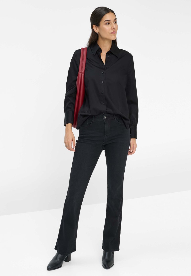 Camicia nera con bottoni, maniche lunghe e colletto, abbinata a jeans scuri. Una borsa a tracolla rossa è appesa a una spalla. Stivali neri alla caviglia.