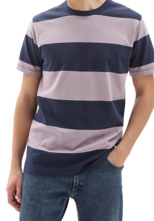 Mand iført kortærmet T-shirt med brede vandrette marineblå og lysegrå striber, kombineret med blå jeans, hånd i lommen.