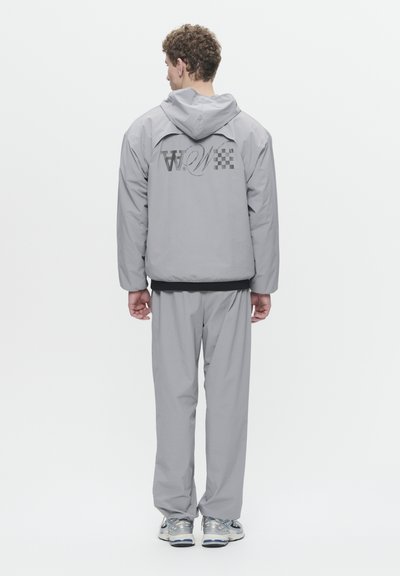 Veste coupe-vent grise avec capuche, arborant un logo et un motif à carreaux au dos. Associée à un pantalon gris assorti. Texture lisse, coupe décontractée.