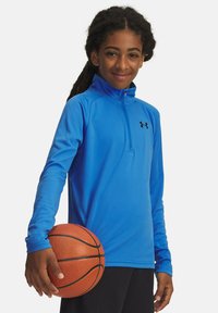Maglietta sportiva blu a maniche lunghe con colletto a mezza zip, con logo nero, indossata da un bambino che tiene un pallone da basket.