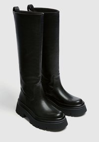 Botas de cuero negras hasta la rodilla con suela de goma gruesa, con un acabado suave, costuras mínimas y lengüetas en la parte superior.