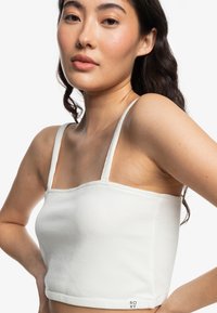 Hvid ribbet cropped top med tynde stropper, med firkantet udskæring og et mærke i nederste side. Glat tekstur og tætsiddende design.