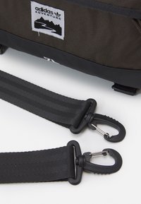 Adventure taška Adidas v tmavě hnědé tkanině s černými akcenty. Obsahuje nylonové popruhy a odolné plastové spony. Zahrnuje nášivku s logem a designem hory.