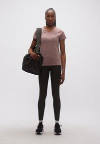 Γυναίκα με πουκάμισο μωβ χρώματος με κοντά μανίκια και μαύρα κολάν στέκεται κρατώντας μια μαύρη αθλητική τσάντα πάνω στον έναν ώμο σε λευκό φόντο.