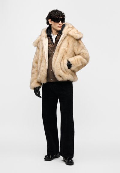 Μπεζ παλτό από συνθετική γούνα με κουκούλα, κλείσιμο με κουμπιά, υφασμάτινη υφή, φορεμένο πάνω από καφέ μπουφάν, συνδυασμένο με μαύρο παντελόνι κοτλέ και γάντια.