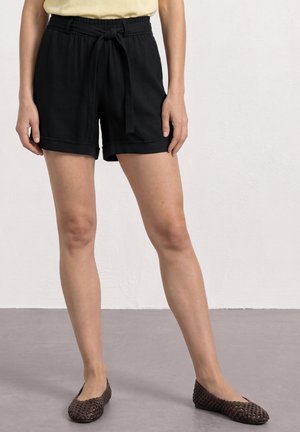 Persoon die zwarte shorts draagt met een gestrikte riem, een lichtgele top en donkerbruine gevlochten platte schoenen, staande op een grijze vloer.