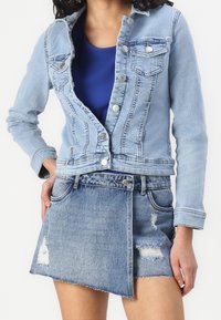 Ljusblå denimjacka med en figurnära design och två bröstfickor, parat med en slitna denim minikjol. Märkbara fransade nederkant detaljer.