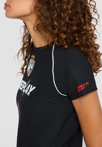 Vrouw met krullend bruin haar, gekleed in een zwart Replay FC sportshirt met logo, witte piping en rode tekst op de mouw, gezien vanaf de zijkant.