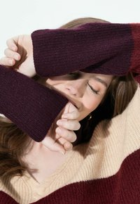 Frau mit geschlossenen Augen, die einen Teil ihres Gesichts mit den Ärmeln eines kastanienbraunen und beigen Strickpullis bedeckt, lächelt sanft vor einem schlichten Hintergrund.