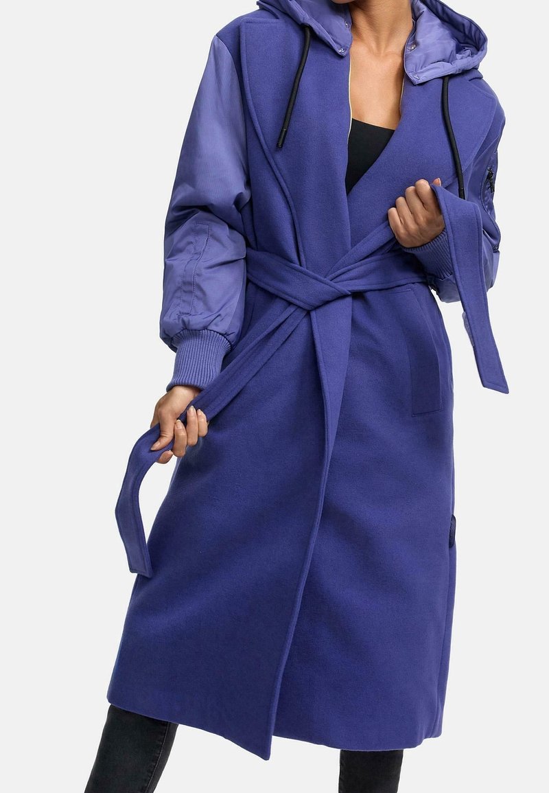 Femme attachant une ceinture sur un long manteau bleu avec des manches matelassées et une capuche, portant un haut noir et un pantalon noir.