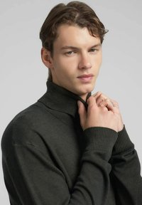 Suéter de cuello alto verde oscuro con puños y cuello acanalados, hecho de una tela suave; el modelo sostiene el cuello en una pose casual.