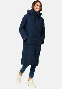 Jūras spēku puffer mētelis ar augstu apkakli un kapuci, ar pilnu priekšējo rāvējslēdzēju un horizontālu quilt dizainu. Apvienots ar ziliem džinsiem un baltām sporta apavām.