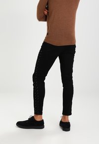 Jeans ajustados negros con detalles de cordones a lo largo de los lados. Combinados con un suéter de punto marrón y zapatos negros. Diseño minimalista y corte ajustado.