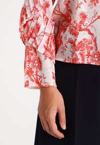 Vit blus med röd blommönster, med volangärmar och textil med struktur. Den visade armen vilar mot en mörk bakgrund.