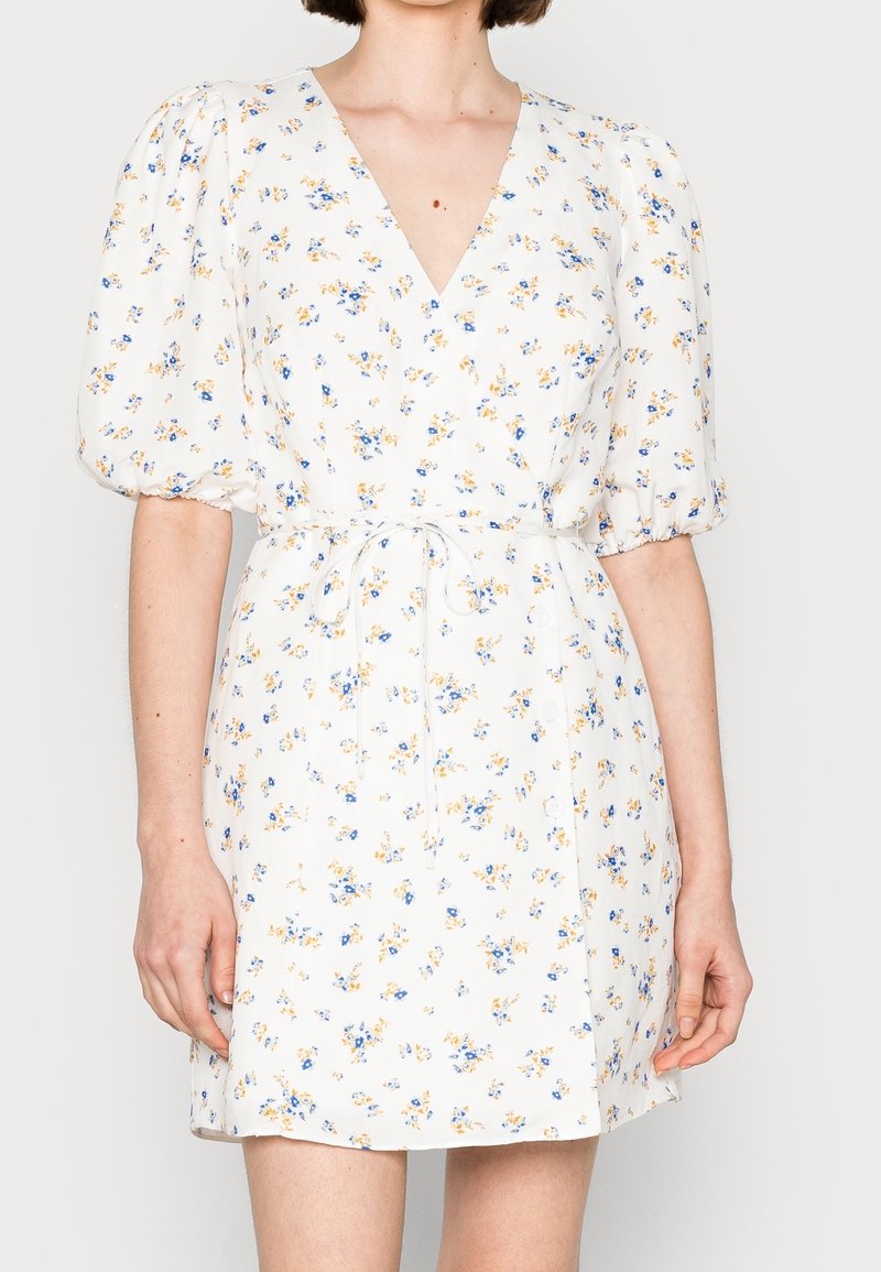 Robe blanche à fleurs avec un décolleté en V profond, des manches bouffantes et une ceinture nouée. Imprimée de petites fleurs bleues et jaunes sur un fond clair.