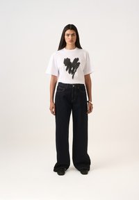 Bílé oversized tričko s grafickým motivem srdce z černého štětce, spárované s tmavě modrými džínami s širokými nohavicemi a černými botami.