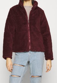 Veste en polaire bordeaux avec col montant et fermeture éclair à l'avant. Présente une surface texturée, deux réglages en bas et des manches longues. Portée sur un jean déchiré.