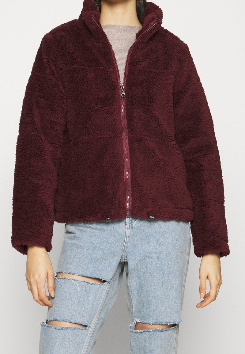 Veste en polaire bordeaux avec col montant et fermeture éclair à l'avant. Présente une surface texturée, deux réglages en bas et des manches longues. Portée sur un jean déchiré.