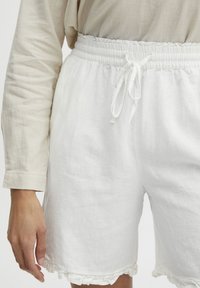 Shorts en lin blancs avec une taille élastique et un cordon de serrage. Présentent des bords froncés à l'ourlet. Texture douce, design décontracté.