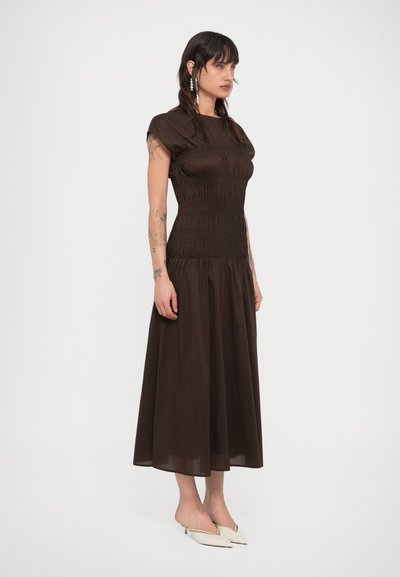 Γυναίκα με τατουάζ που φοράει midi φόρεμα σε σκουρό καφέ με ρυτιδώσεις και λευκά μυτερά τακούνια, στέκεται μπροστά σε απλό λευκό φόντο.